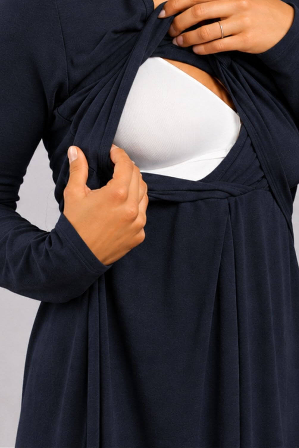 A close-up of a woman wearing a navy blue long-sleeve maternity cover-up dress over a white bra, demonstrating how to open the front closure with her hands