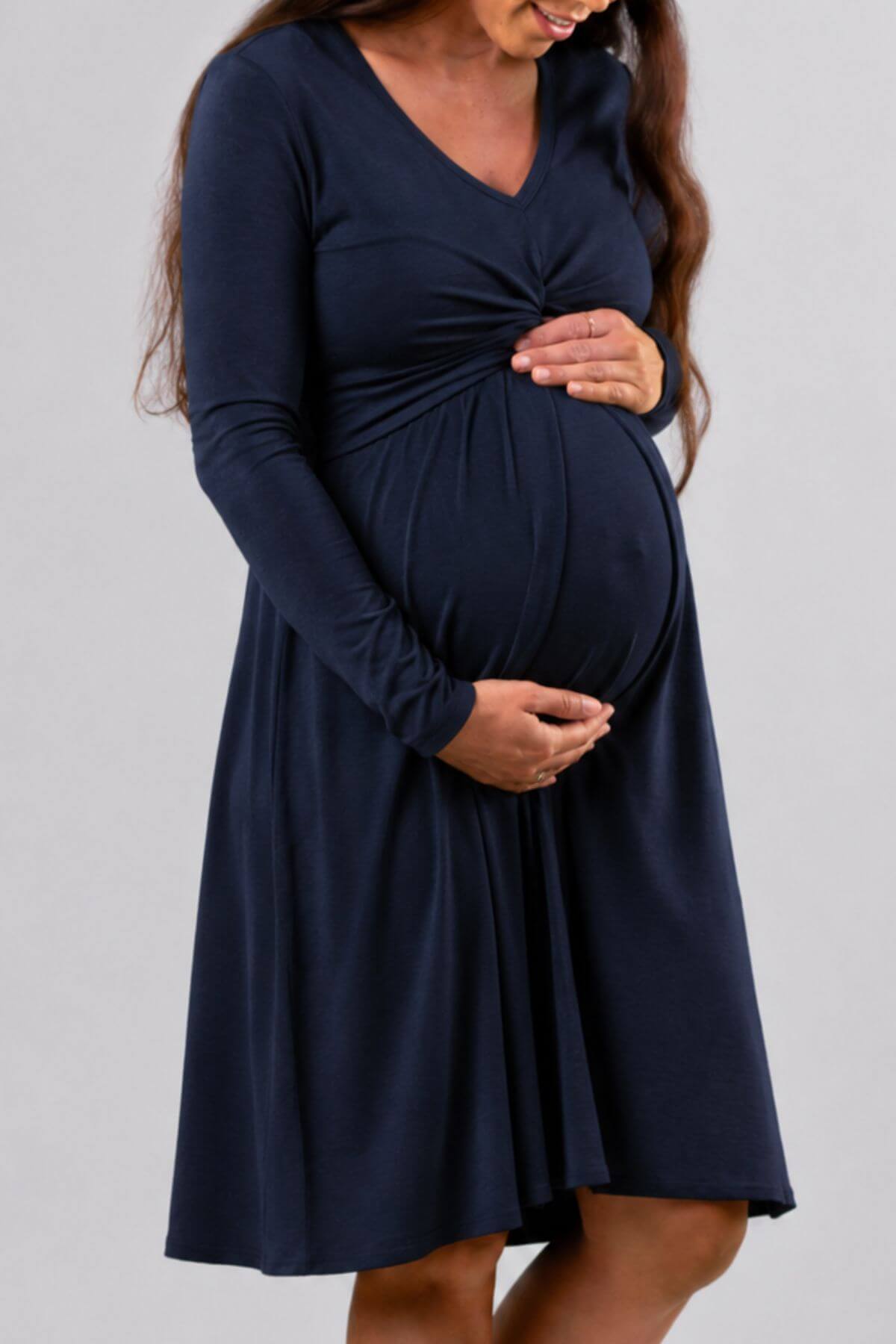 A pregnant woman wearing a long-sleeve navy blue maternity dress with a V-neck and gathered waist, standing against a solid light gray background