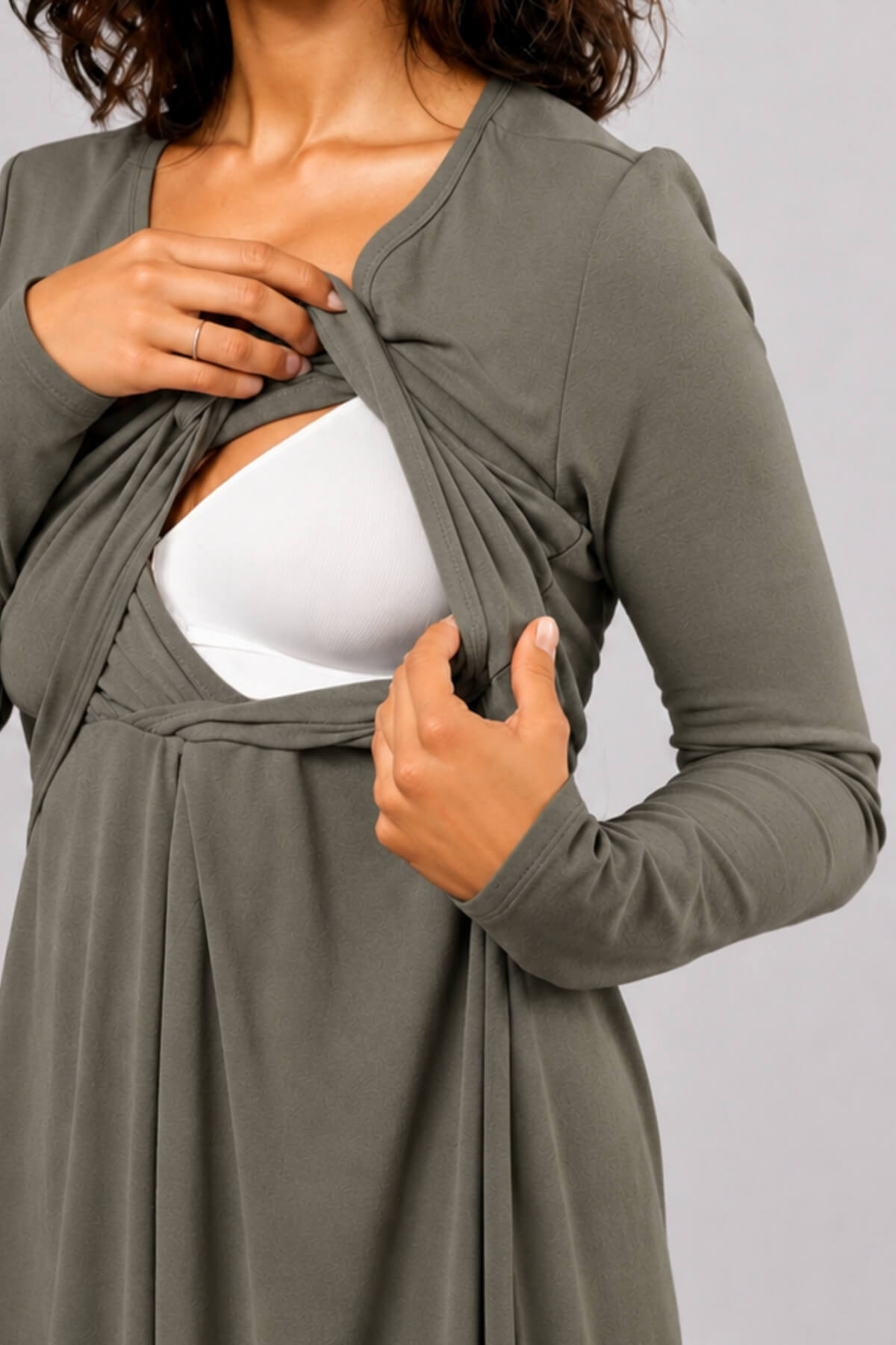 A close-up of a woman wearing an olive green long-sleeve wrap dress or cardigan over a white bralette, with her hands lifting the fabric to reveal the undergarment. The background is a solid light gray studio backdrop