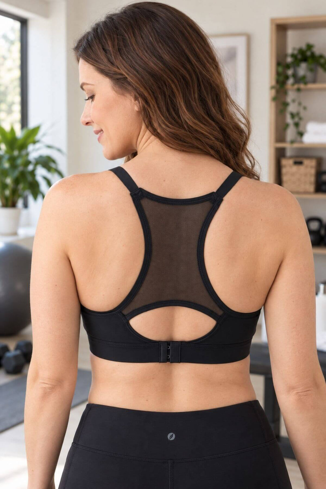 A woman shown from the back wearing a black Simple Wishes nursing and hands free pumping sports bra with a sheer mesh back panel and keyhole cutout, paired with black leggings in a bright indoor room.