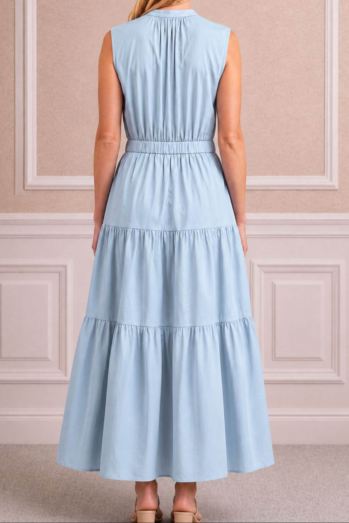 A woman viewed from the back wearing a sleeveless light blue tiered maxi dress with a gathered waist, standing in front of a beige paneled wall.