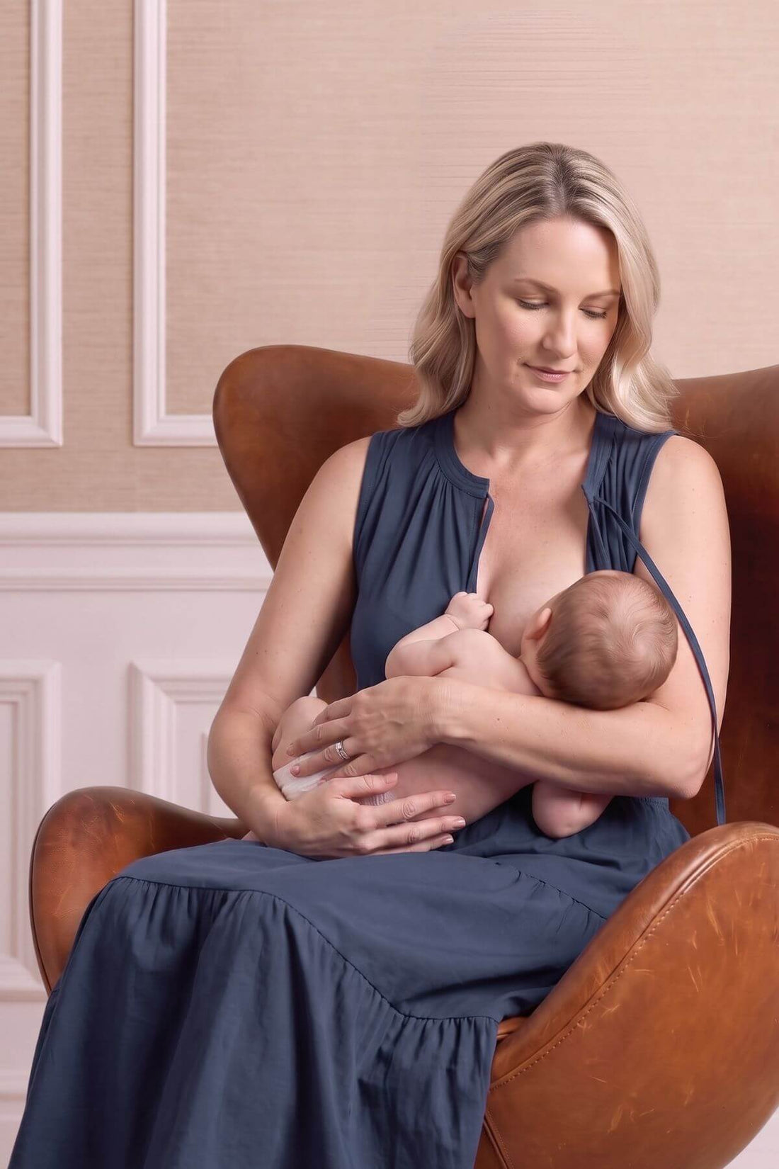 A woman in a navy blue maternity dress breastfeeding a baby while seated in a brown leather armchair against a beige paneled wall.