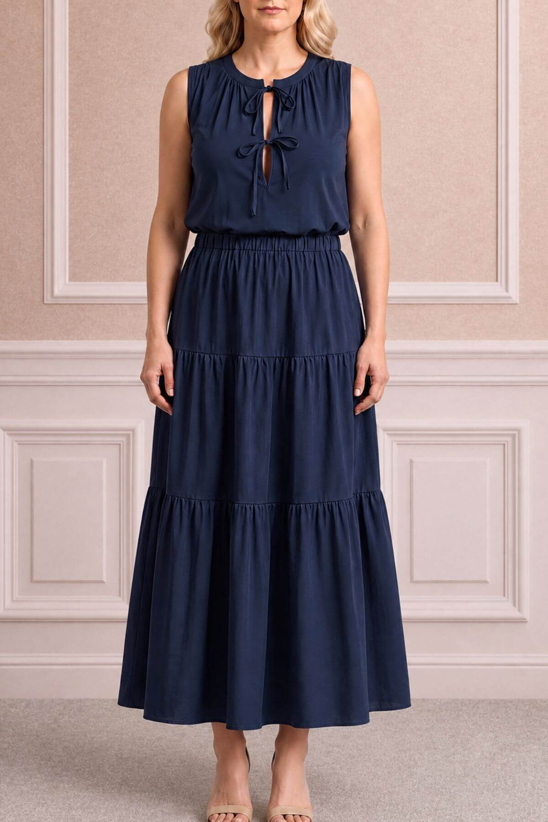 A woman modeling a sleeveless navy blue maxi dress with a tiered skirt and a keyhole tie-neck detail, standing against a beige paneled wall.