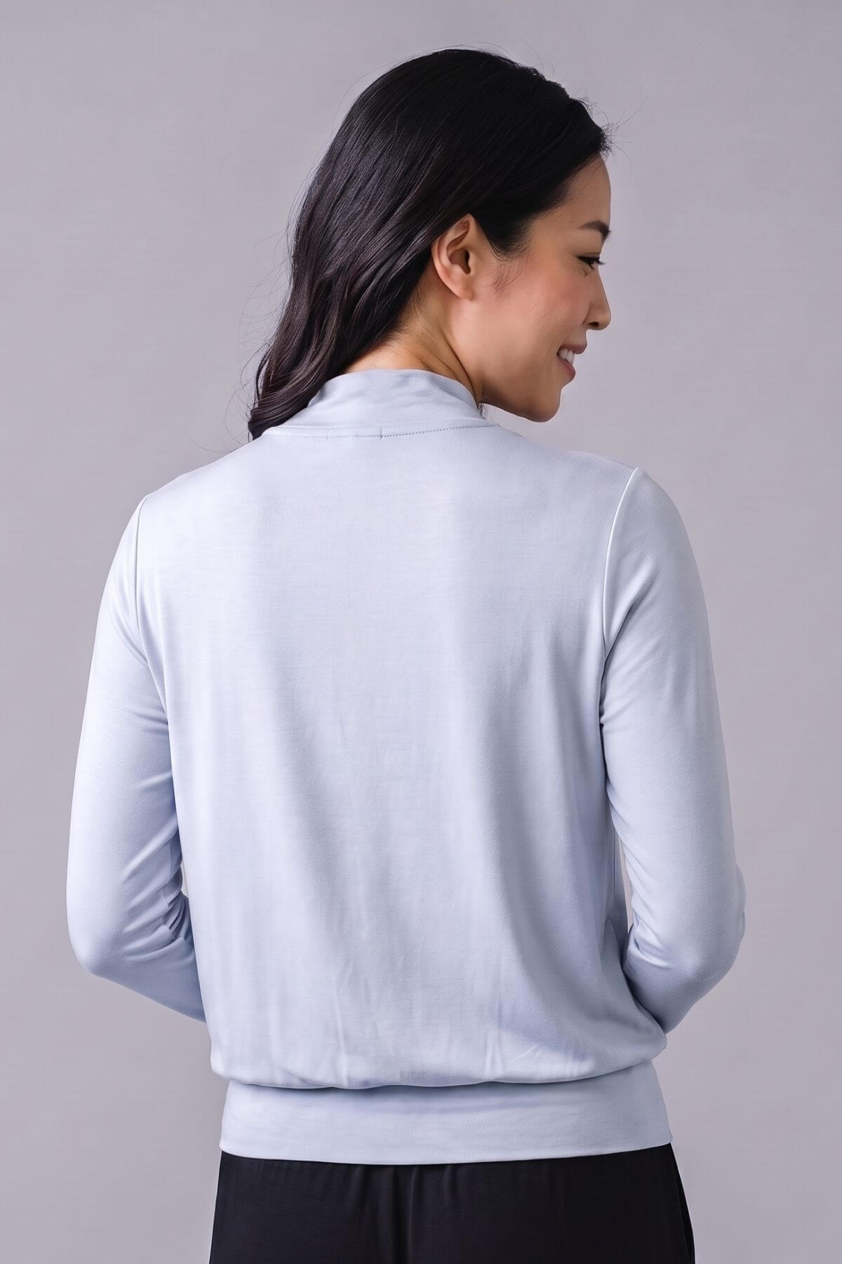 A woman shown from the back wearing a light blue long-sleeve knit top with a mock neck and ribbed hem, paired with black bottoms, against a solid gray studio background.