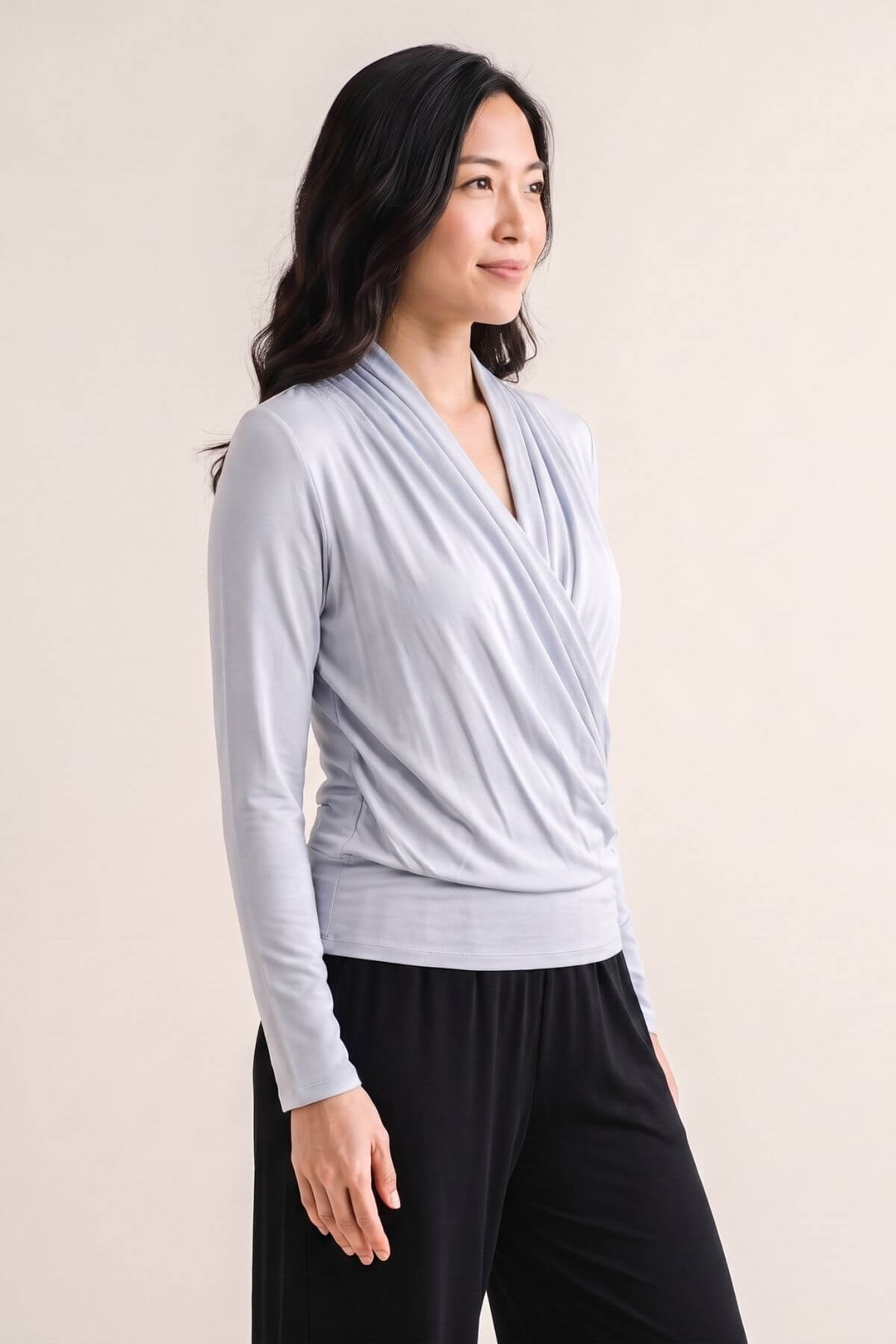 A woman wearing a light gray long-sleeve wrap-style top and black pants, standing against a plain beige studio background.