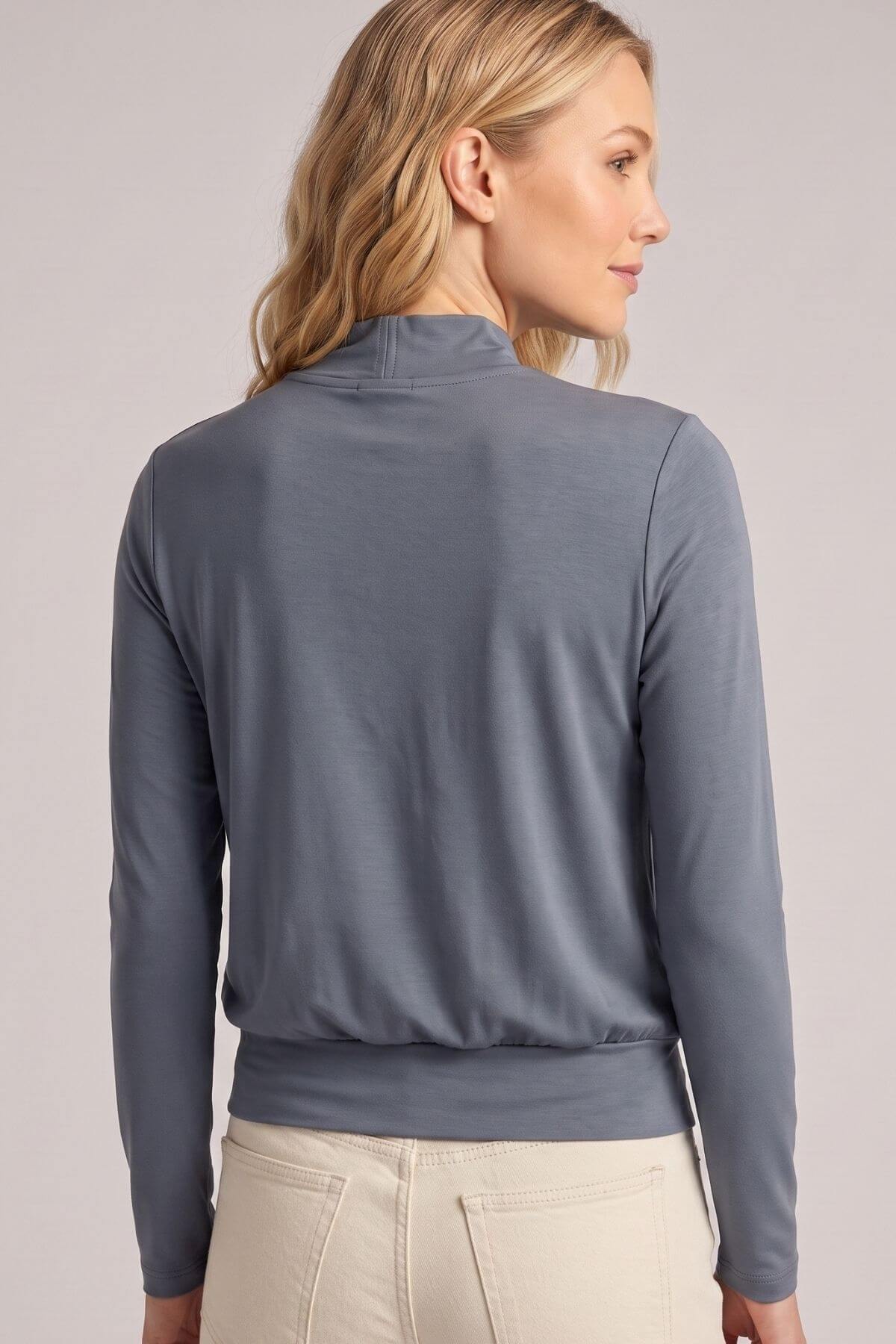 A woman shown from the back wearing a slate blue-gray mock neck long-sleeve top and light beige pants, against a neutral light gray studio background.
