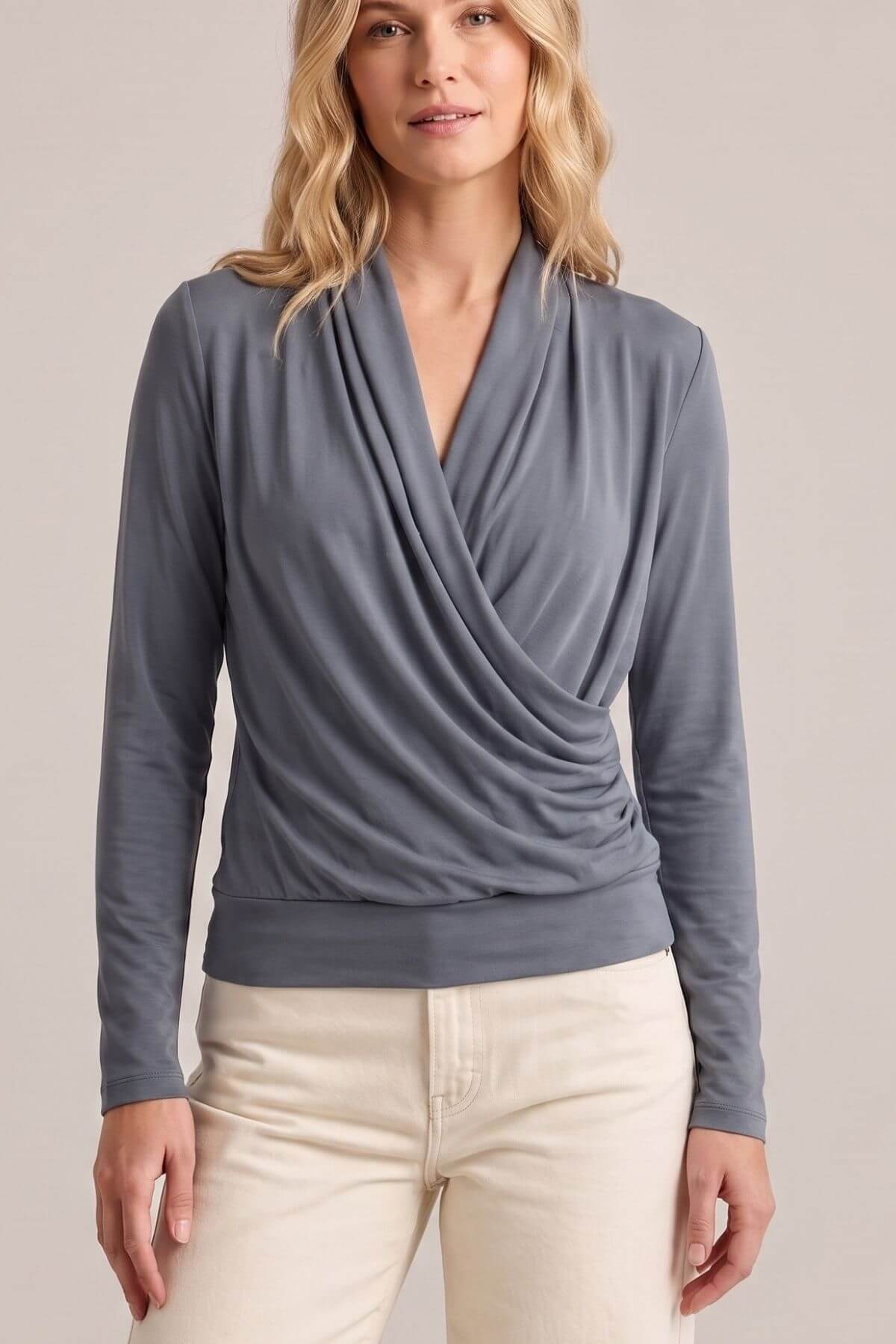 A woman wearing a slate blue-gray long-sleeve wrap-style top with a draped front, paired with cream pants, against a plain light beige studio background.