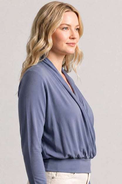 A woman with blonde hair wearing a slate blue long-sleeve wrap top, shown from a side profile against a light gray studio background.
