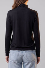 A woman shown from the back wearing a black long-sleeve mock neck top and light blue jeans against a plain light gray studio background.d.#color_midnight