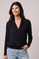 A woman wearing a black long-sleeve wrap-style top with a draped V-neck, paired with light blue jeans, photographed against a plain light gray studio background.#color_midnight