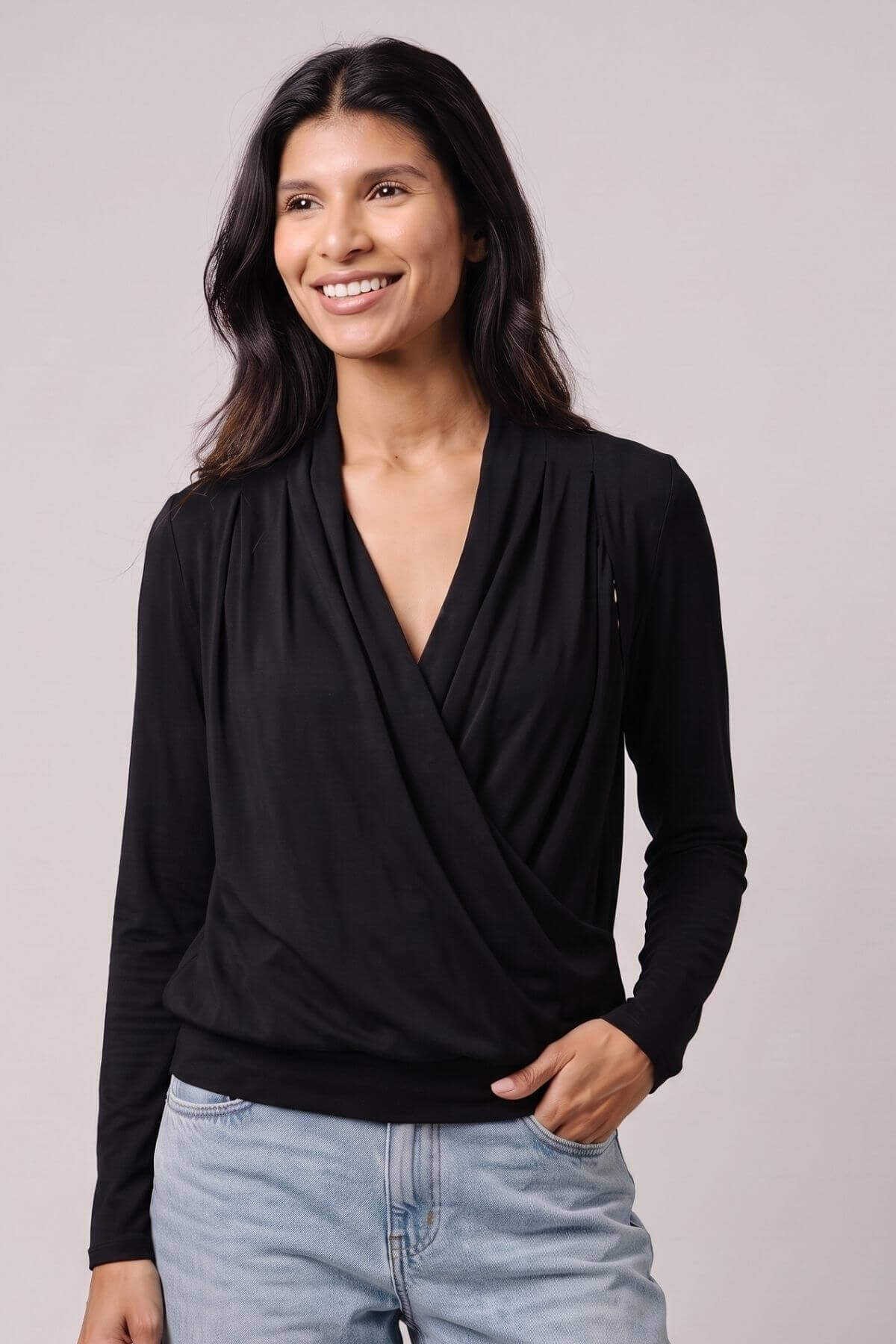 A woman wearing a black long-sleeve wrap-style top with a draped V-neck, paired with light blue jeans, photographed against a plain light gray studio background.