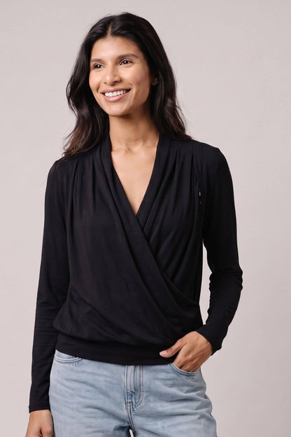 A woman wearing a black long-sleeve wrap-style top with a draped V-neck, paired with light blue jeans, photographed against a plain light gray studio background.