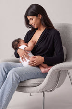 A woman in a black long-sleeve top and light blue jeans sits in a light gray chair, breastfeeding a baby dressed in a white onesie. The background is a clean, neutral studio setting.d.#color_midnight