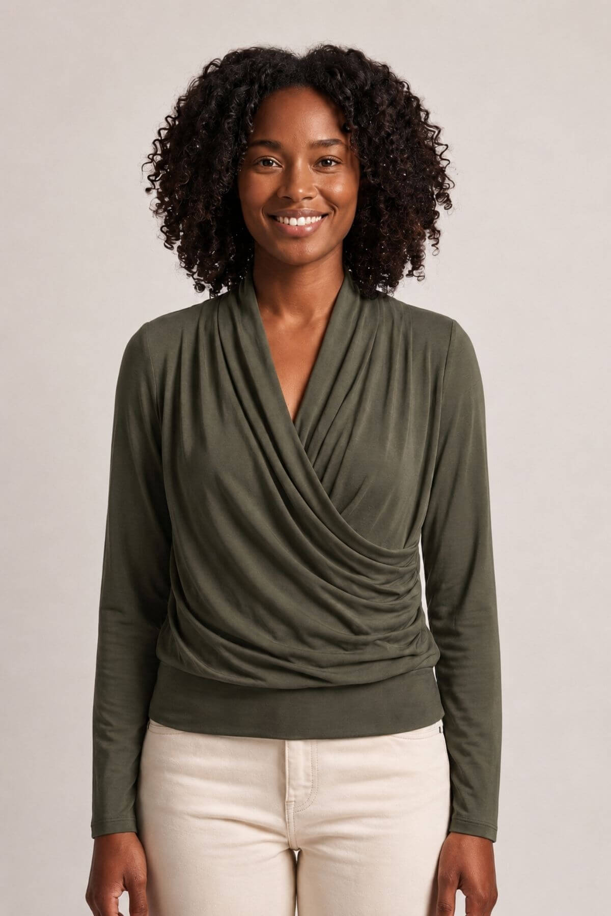 A smiling woman with curly black hair wearing an olive green long-sleeve draped wrap top and cream-colored pants, standing against a light gray studio background.