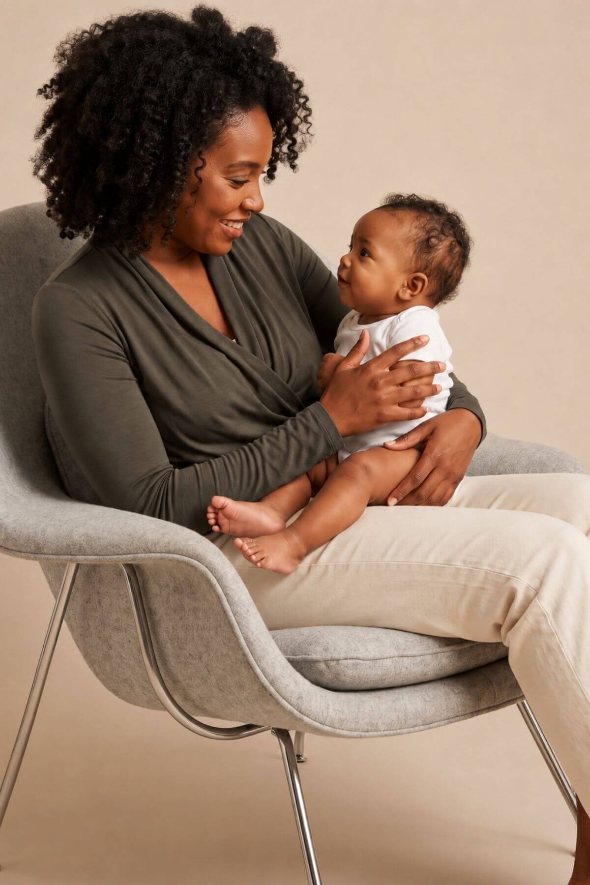 A woman with curly black hair sits in a light gray upholstered chair, holding a baby. The woman wears an olive green long-sleeve top and beige pants, while the baby is dressed in a white outfit.