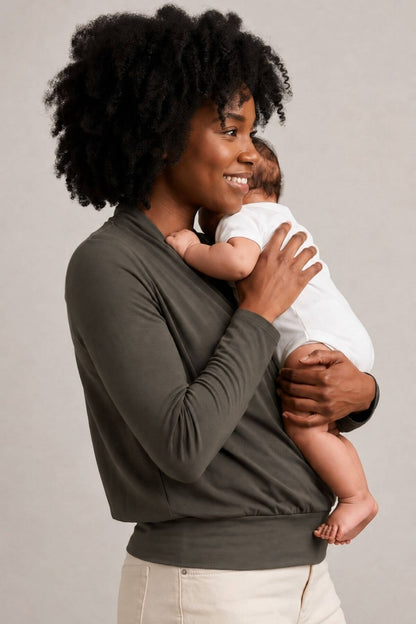 A woman with dark curly hair holds a baby wearing a white onesie. She is wearing a dark olive green long-sleeve top and light beige pants, photographed against a neutral light gray studio background.