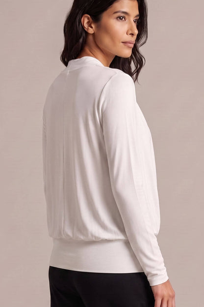 A woman shown from the back wearing a white long-sleeve knit top and black pants, photographed against a plain warm beige studio background.
