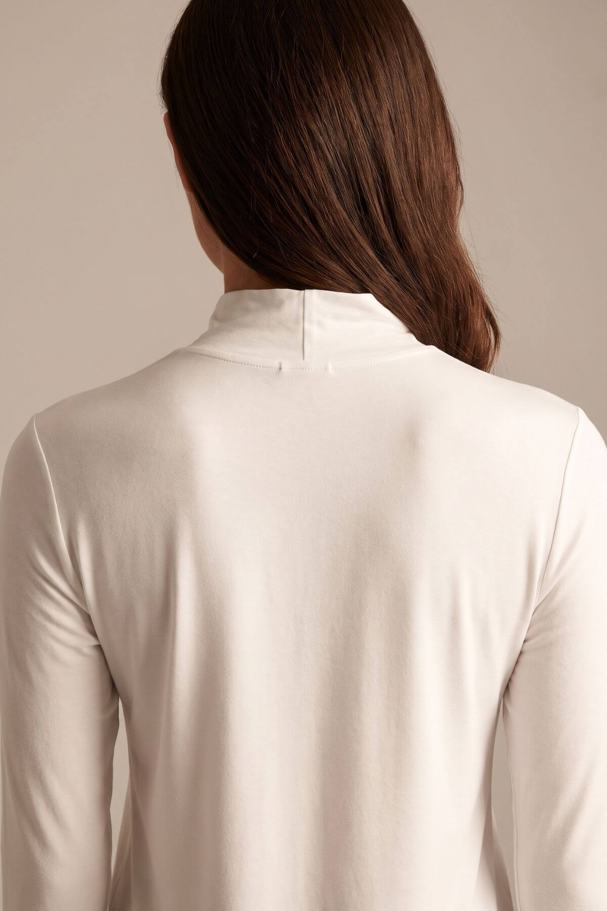 Back view of a woman with long brown hair wearing a cream-colored long-sleeve mock neck top against a solid beige background..