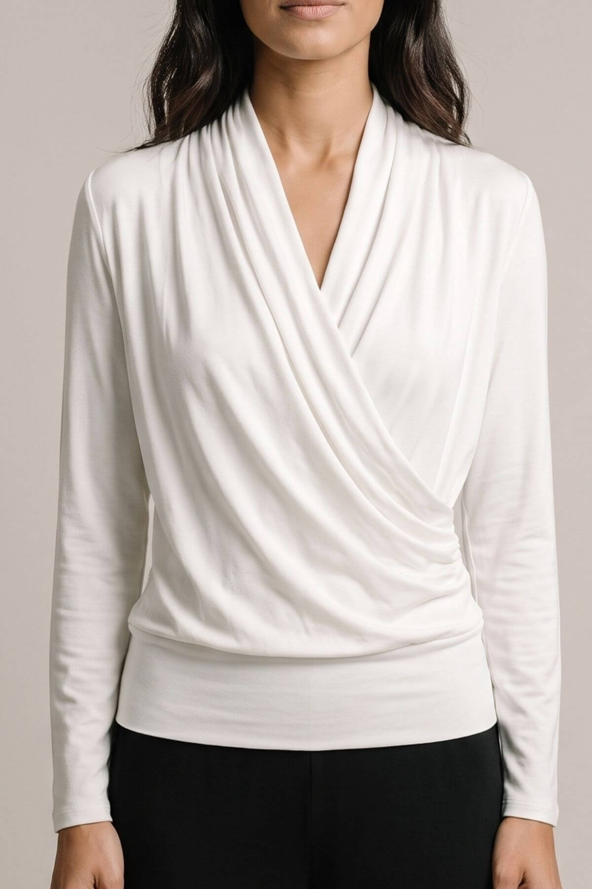 A woman wearing a white long-sleeve wrap-style top with a draped crossover front and a wide ribbed hem, paired with black bottoms, against a plain light beige studio background..