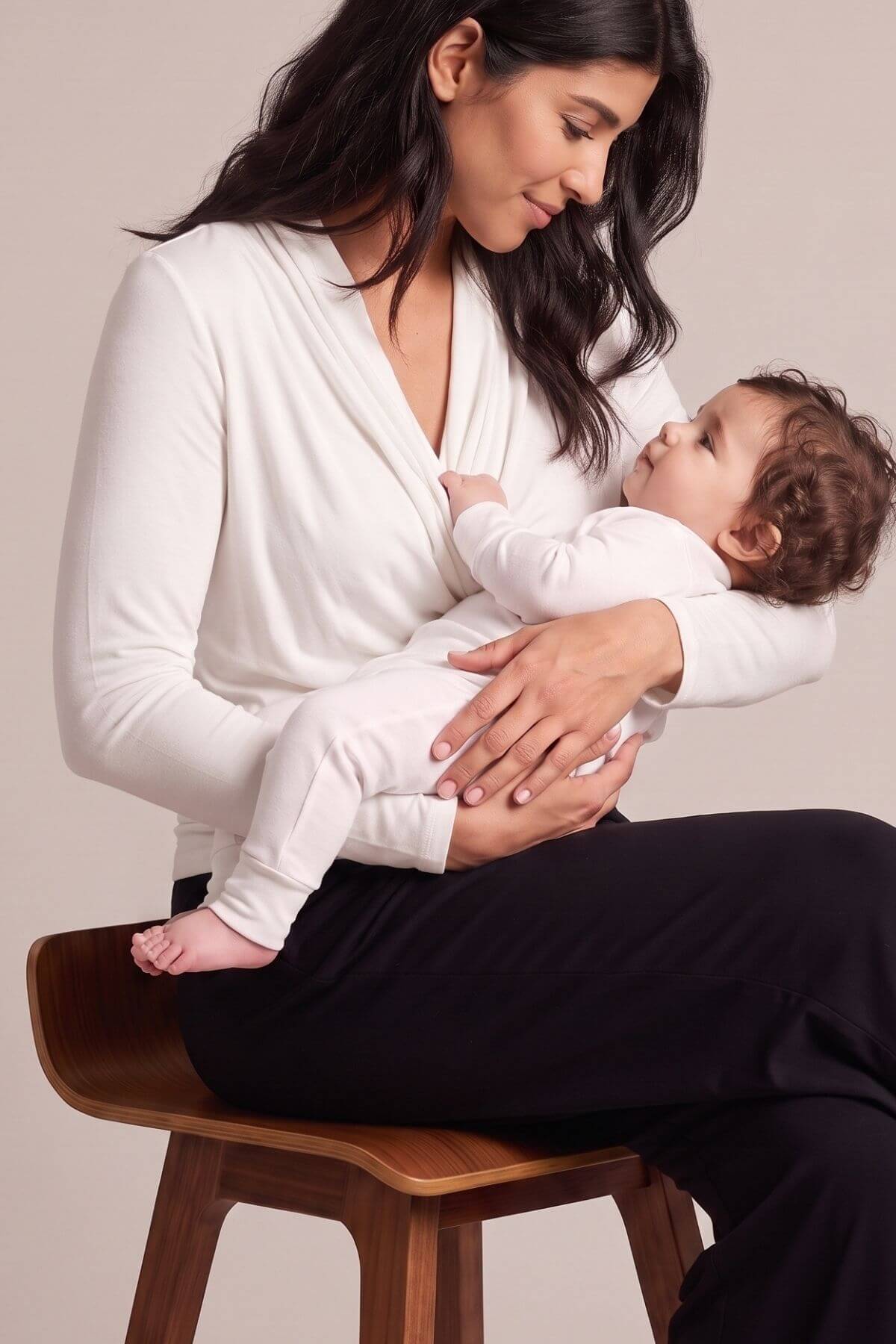 A woman sitting on a wooden stool holds a baby wearing a white long-sleeve bodysuit. The woman wears a white V-neck long-sleeve top and black pants against a plain beige studio background.