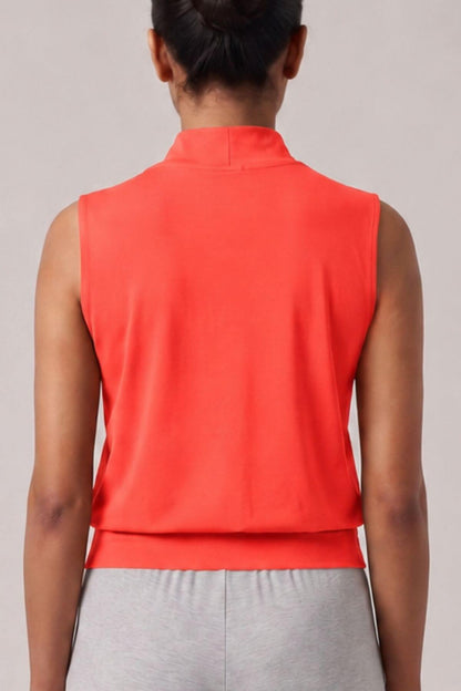 A woman viewed from the back wearing a sleeveless high-neck top in a bright coral-orange color, paired with light gray sweatpants, against a plain light gray studio background.