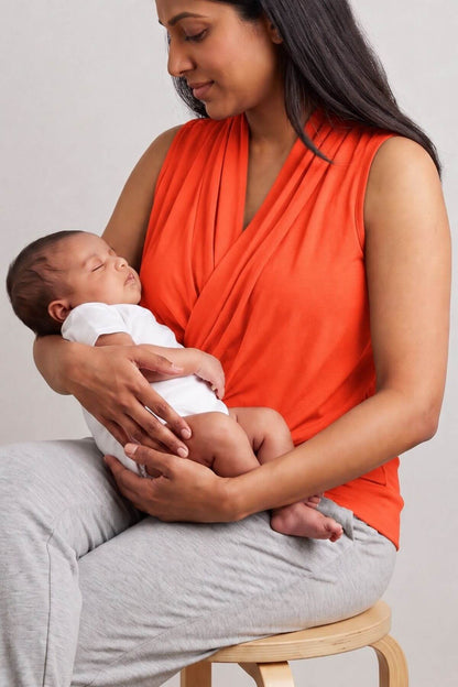 A woman wearing a bright orange draped wrap-style top sits on a wooden stool and holds a sleeping newborn baby dressed in a white outfit. The background is a clean, neutral light gray studio wall.