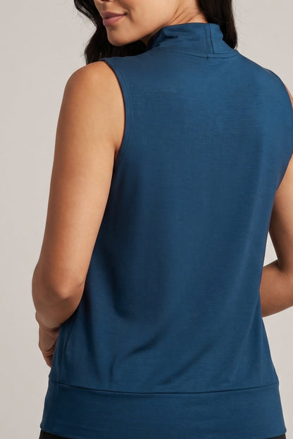 A close-up back view of a woman wearing a sleeveless mock neck athletic top in a solid medium blue color, set against a plain light gray studio background.