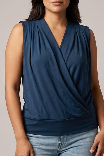 A woman wearing a sleeveless navy blue wrap-style top with a draped V-neckline, paired with light blue jeans, against a plain light gray studio background.