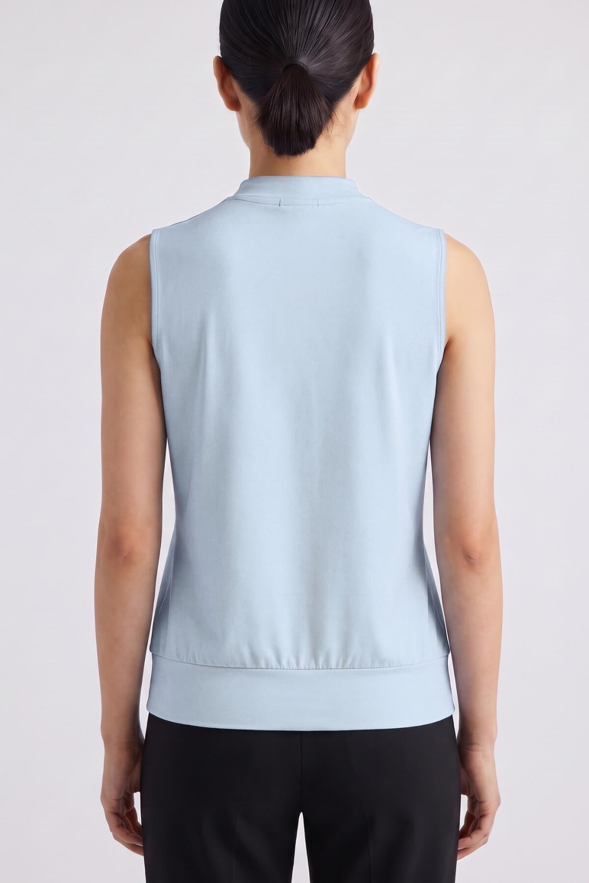 A woman viewed from the back wearing a sleeveless light blue mock-neck top and black pants against a plain light gray studio background.