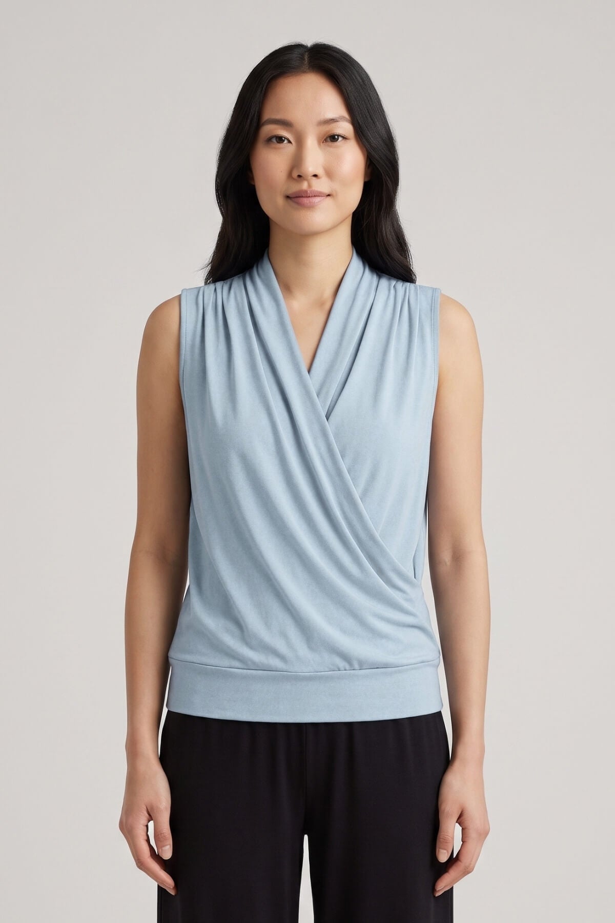 A woman modeling a light blue sleeveless wrap-style top with a draped V-neckline, paired with black pants, against a plain light gray studio background.