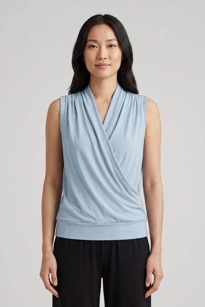 A woman modeling a light blue sleeveless wrap-style top with a draped V-neckline, paired with black pants, against a plain light gray studio background.