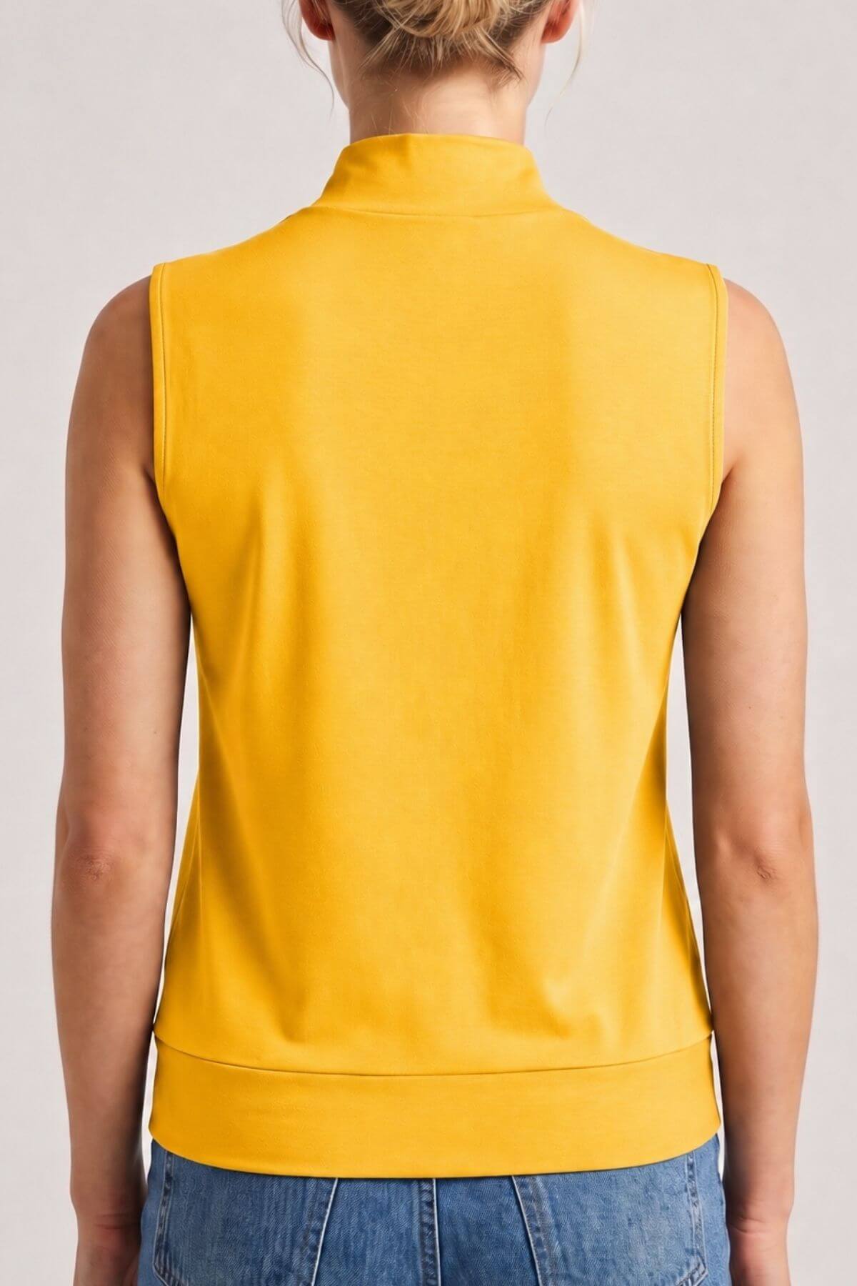 A woman viewed from the back wearing a sleeveless mustard yellow mock-neck top and blue jeans against a light gray studio background..