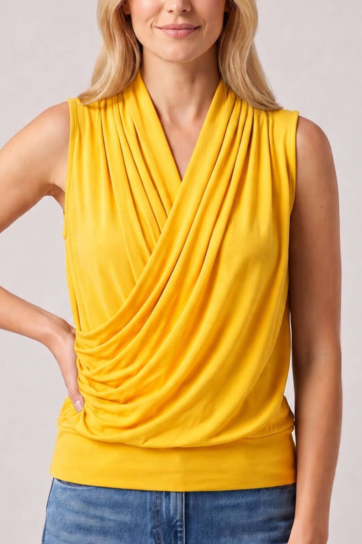 A woman wearing a bright yellow sleeveless draped top with a crossover V-neck, paired with blue jeans, against a light gray studio background.