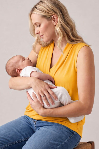 A woman in a yellow sleeveless top and blue jeans holds a baby dressed in a white onesie while breastfeeding. The photo is shot in a studio setting against a plain light gray background..