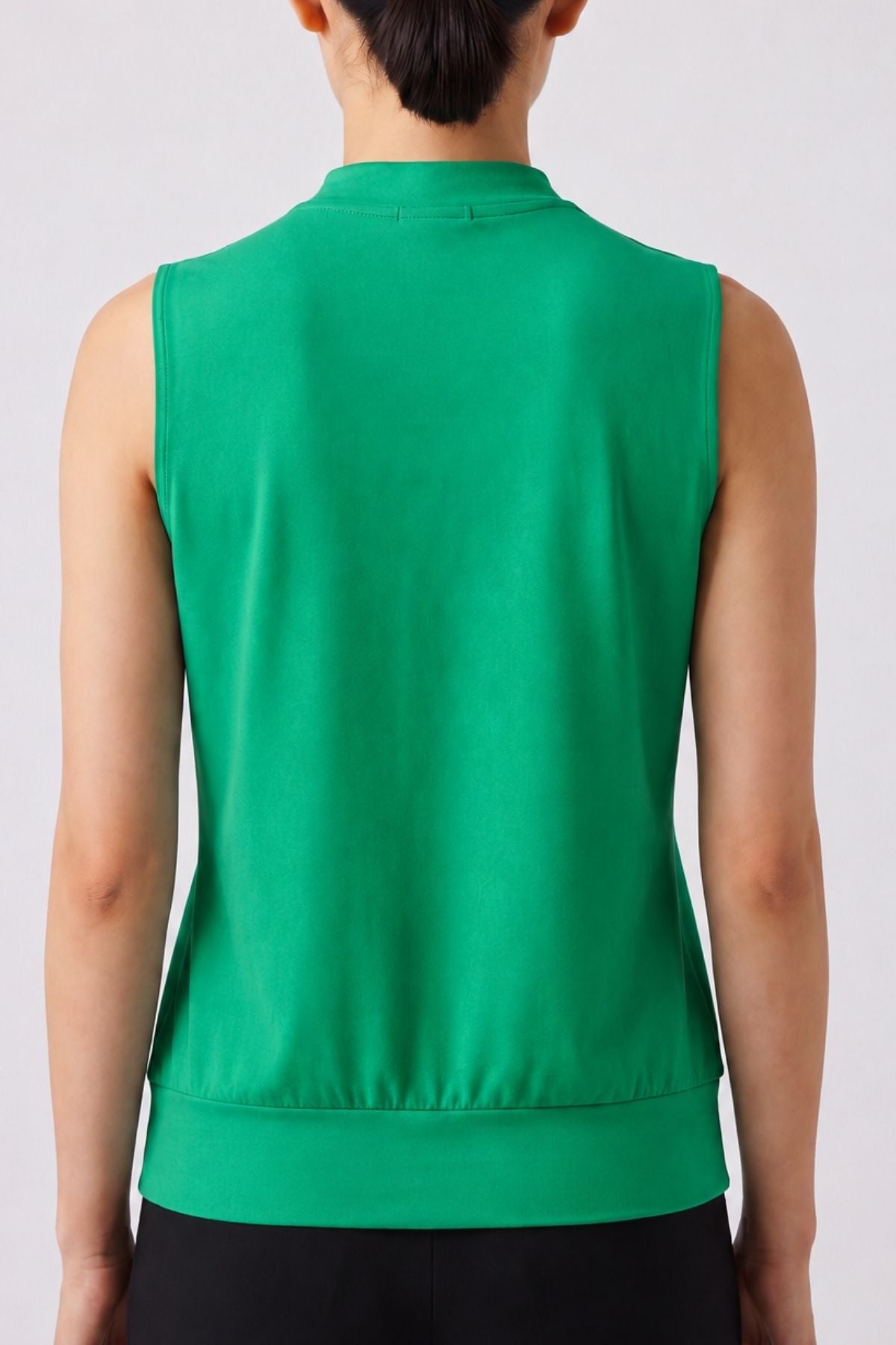 A person shown from the back wearing a sleeveless green top with a high mock neck and ribbed hem, paired with black bottoms, against a light gray studio background.