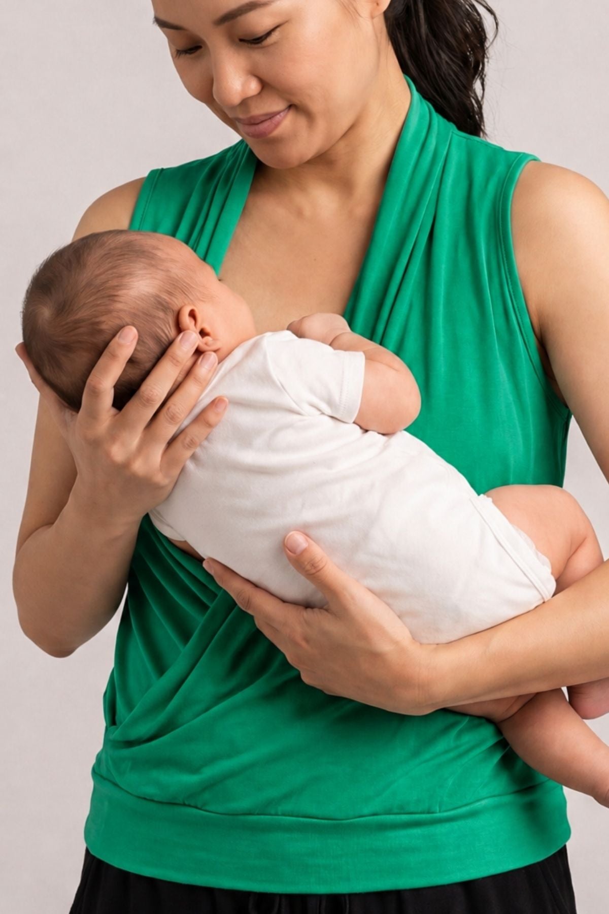 A woman wearing a bright green sleeveless baby wrap carrier holds a baby dressed in a white onesie against a plain light gray background.
