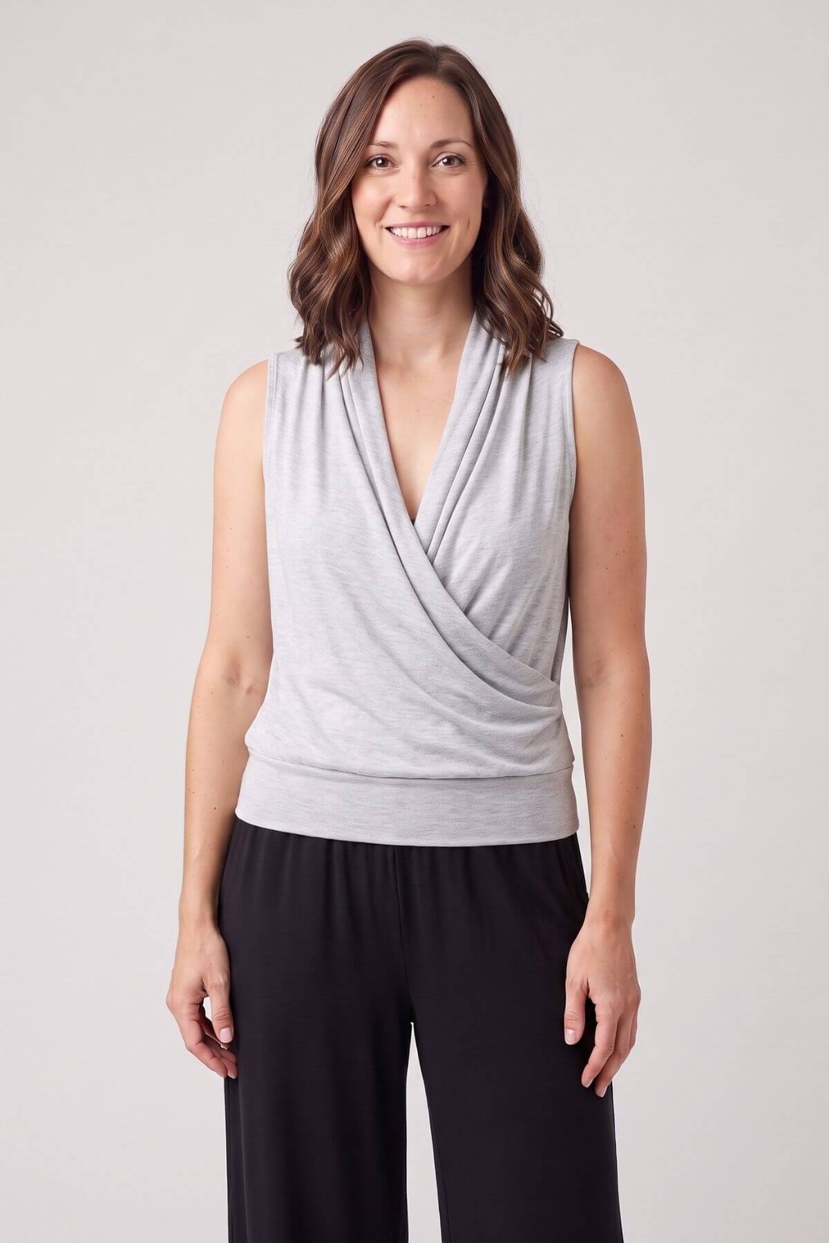 A woman modeling a light gray sleeveless wrap-style top with a draped crossover front, paired with black pants, against a plain light gray studio background.