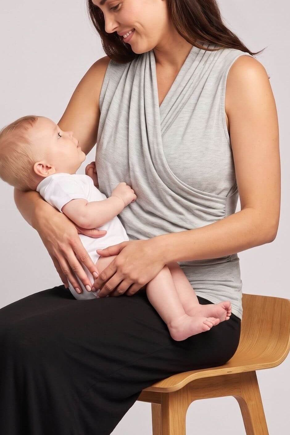 A woman wearing a light gray draped sleeveless top and black pants sits on a wooden stool, holding a baby dressed in a white outfit. The background is a clean, neutral light gray studio backdrop.