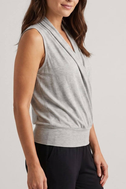 A woman wearing a light gray sleeveless wrap-style top and dark pants, photographed against a plain light gray studio background.