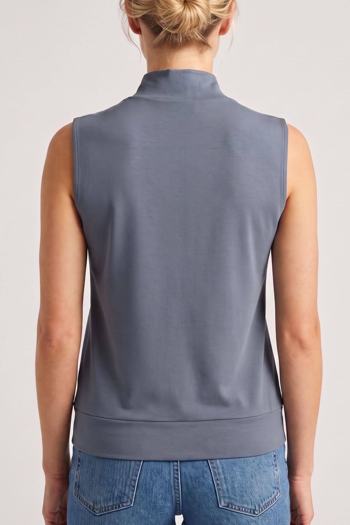 A woman viewed from the back wearing a sleeveless mock-neck top in a muted slate blue-gray color, paired with blue jeans, against a plain light gray studio background.