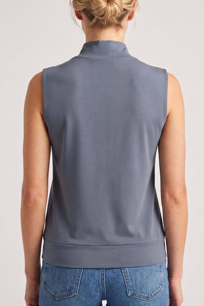 A woman viewed from the back wearing a sleeveless mock-neck top in a muted slate blue-gray color, paired with blue jeans, against a plain light gray studio background.