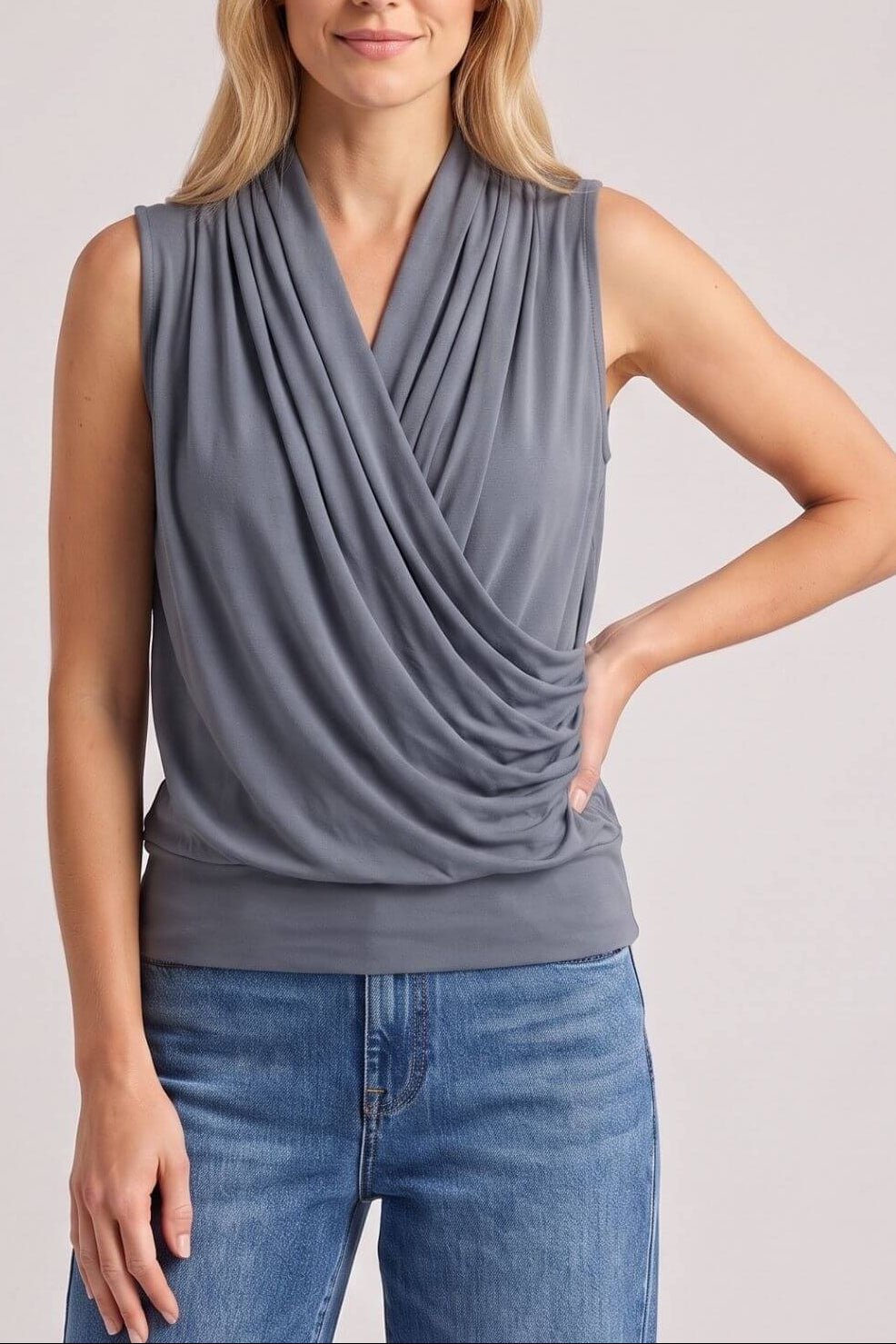 A woman modeling a sleeveless draped wrap-style top in a medium gray color, paired with blue jeans, against a solid light gray studio background.