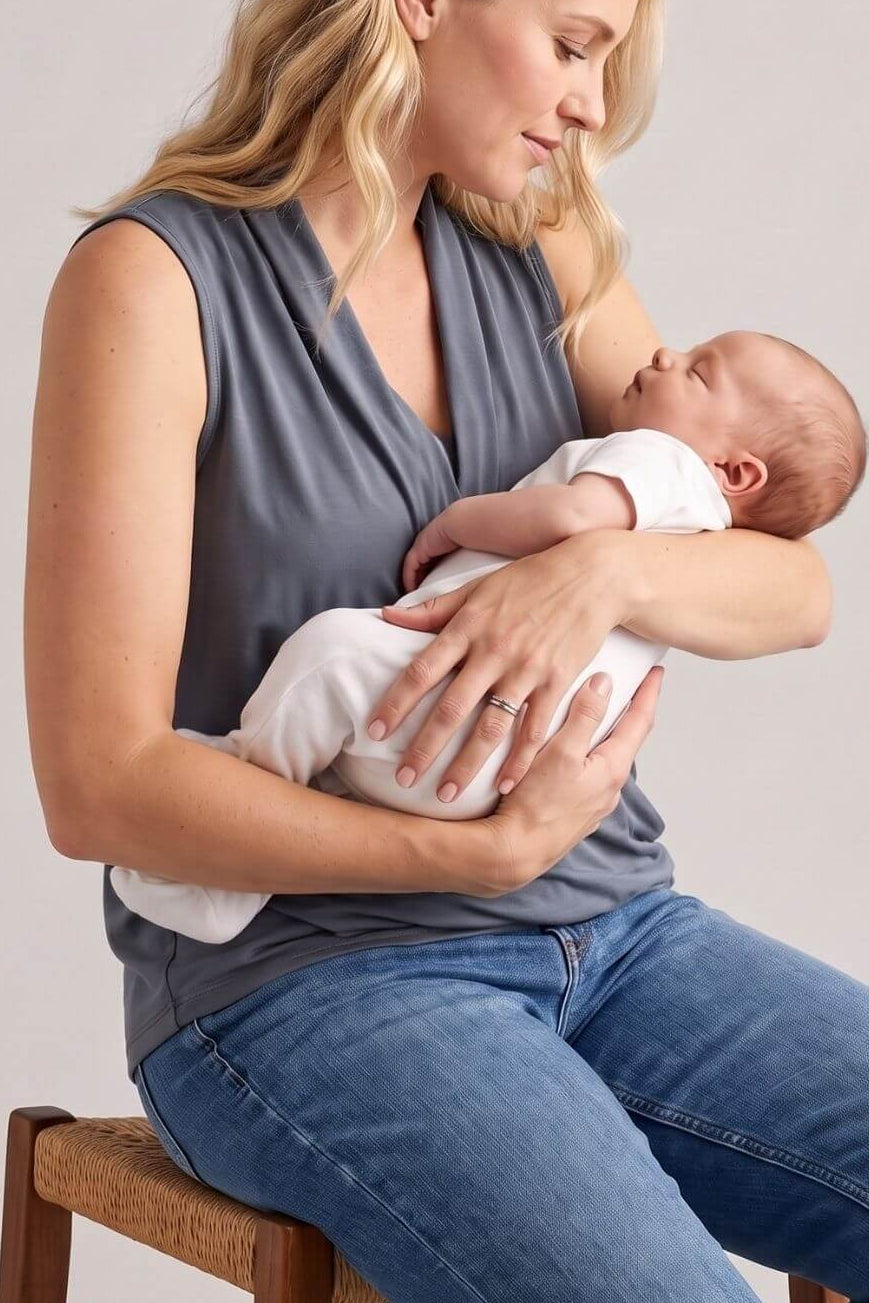 A woman wearing a gray sleeveless top and blue jeans sits on a wooden stool, breastfeeding a baby wrapped in a white blanket. The photo is shot in a studio setting with a plain light gray background.