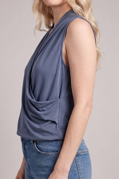 A woman wearing a sleeveless draped wrap-style top in a muted slate blue color, paired with blue jeans, shown from a side profile against a plain light gray studio background.