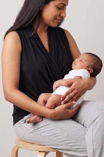 A woman wearing a black sleeveless wrap top sits on a wooden stool, holding a sleeping baby dressed in a white outfit. The background is a clean, neutral light gray studio wall.