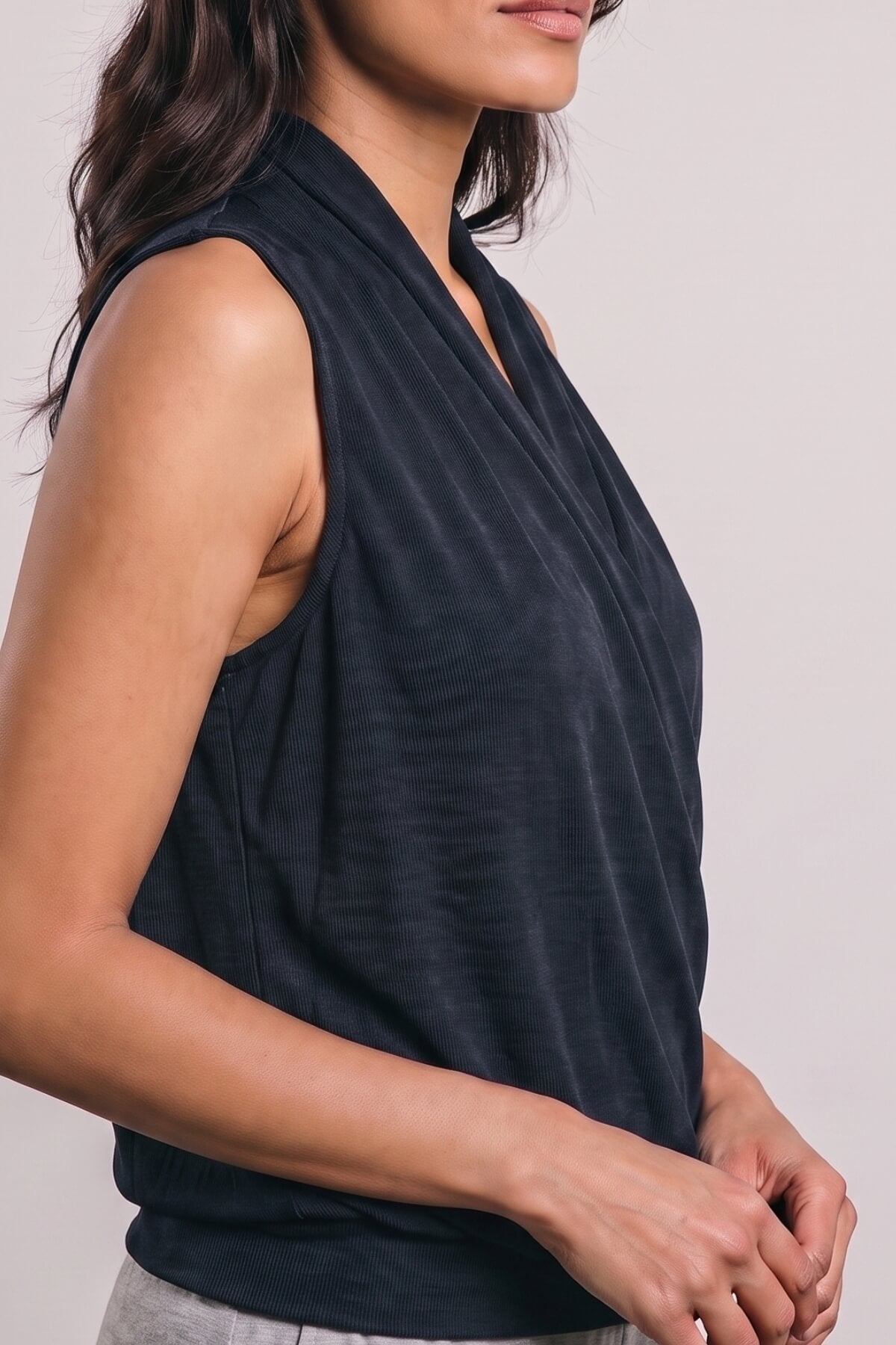 A close-up side view of a woman wearing a sleeveless, draped V-neck top in a dark charcoal gray color, paired with light gray bottoms, against a plain light gray background.