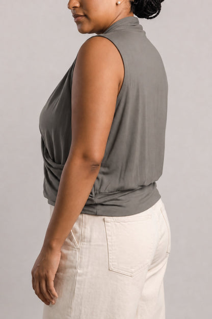 A woman shown from the side wearing a sleeveless, high-neck, wrap-style top in a muted gray-green color, paired with off-white pants, against a plain light gray studio background.