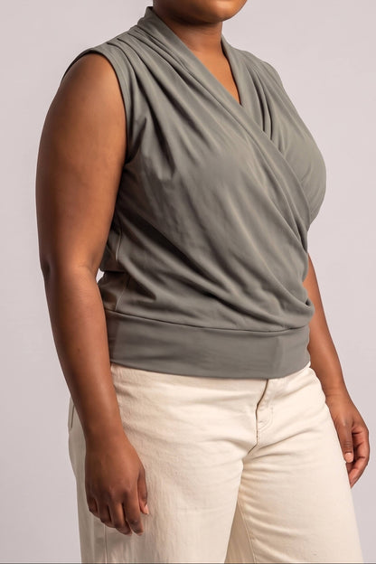A woman wearing a sleeveless draped wrap-style top in a muted sage green, paired with light cream pants, against a plain light gray studio background.