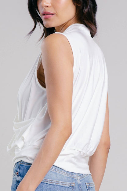 A woman wearing a white sleeveless draped top and light blue jeans, shown from a side-back angle against a light gray studio background.