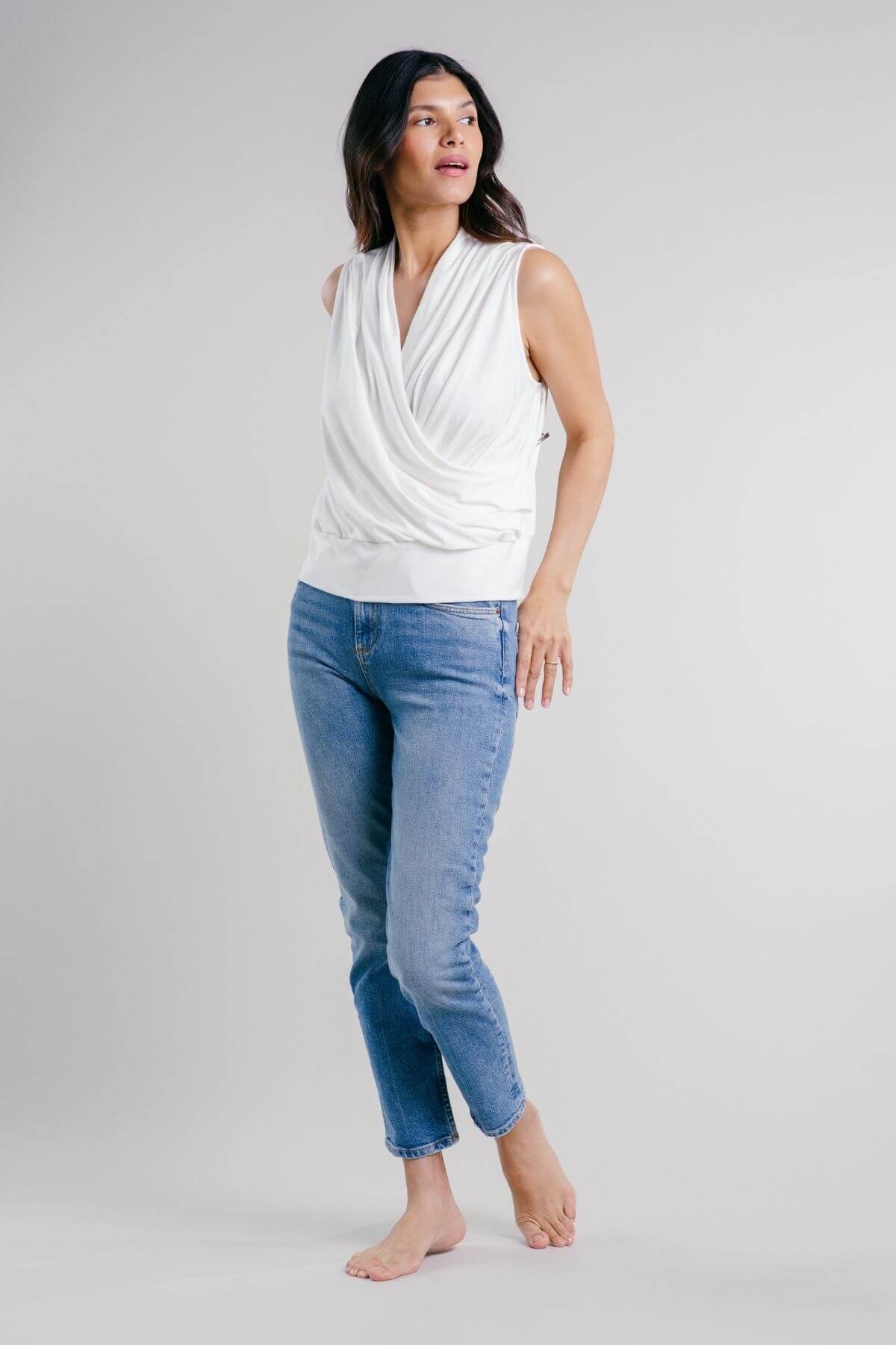 A woman modeling a white draped sleeveless top and light blue cropped jeans against a plain light gray studio background.